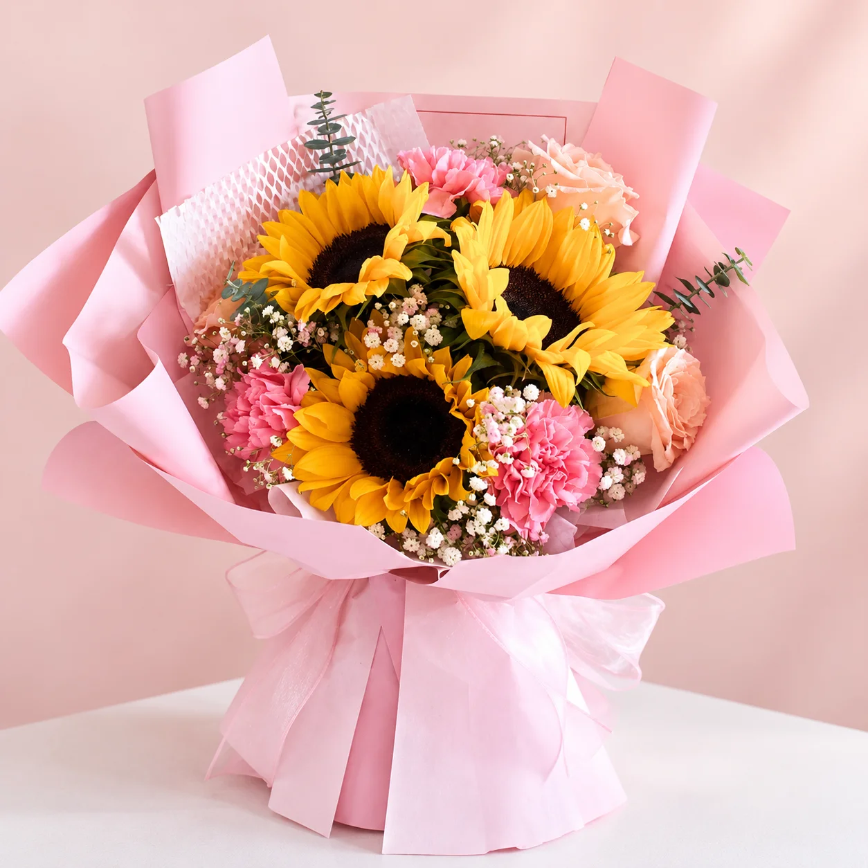 Girasoles Rosé - Ramo de 3 girasoles con claveles rosados, rosas durazno, flor nube y eucalipto, envuelto en papel rosa con lazo